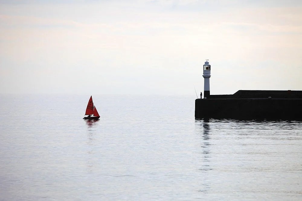 Red-sailed boat passing a harbour lighthouse on a still, misty morning in Cornwall