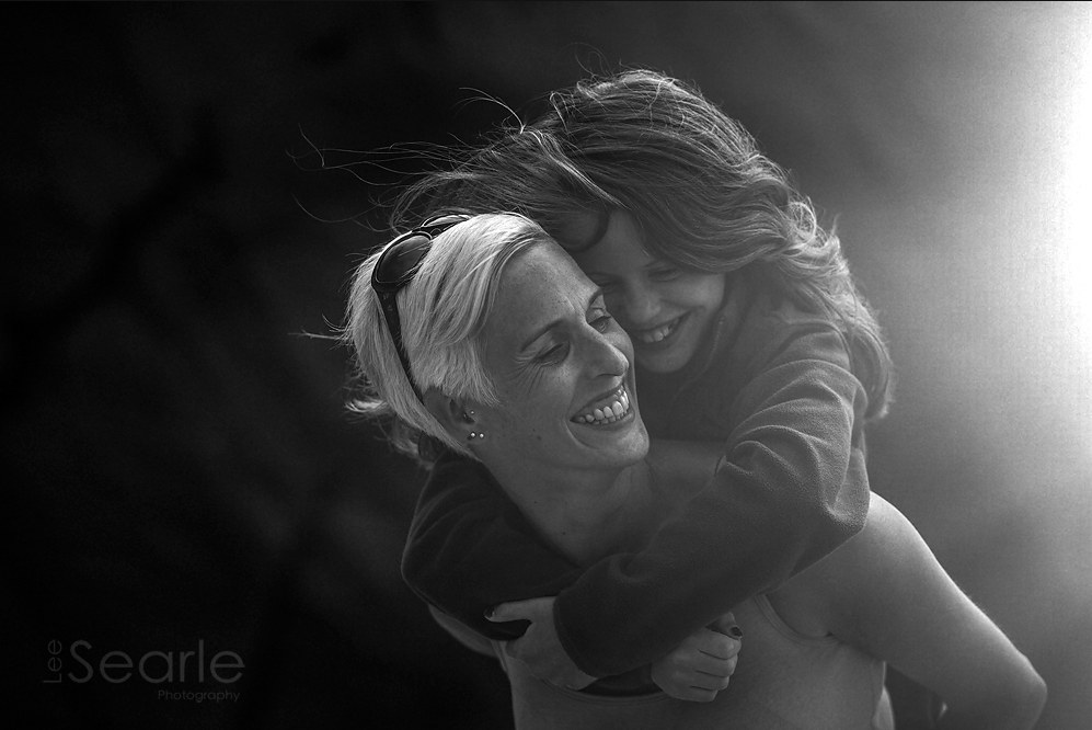 Black and white photograph of a mother giving her daughter a piggyback, laughing together in Cornwall
