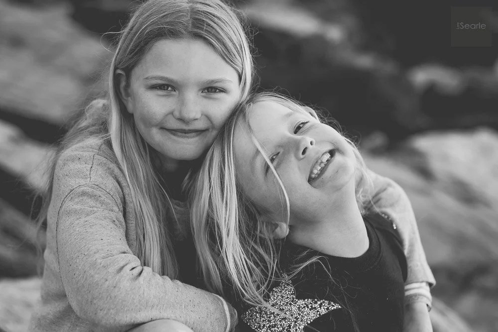 Black and white portrait of two sisters leaning together and laughing on the Cornish coast