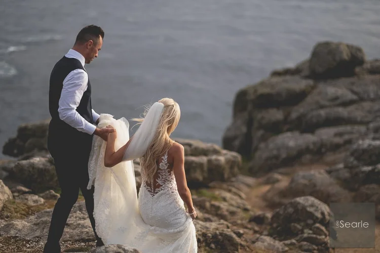 Groom carrying the veil as the bride picks her way down the cliff path