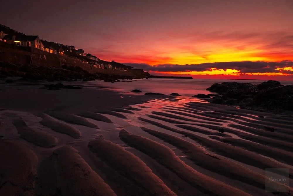 Rippled sand glowing under a blazing sunset over a Cornish fishing village